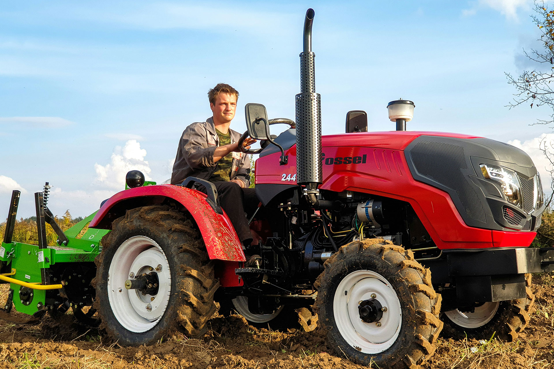 Kompakttraktoren eignen sich ideal für Arbeiten auf engem Raum und sind auch gebraucht eine attraktive Alternative. Junger Mann fährt einen roten Kleintraktor mit angebauter grüner Bodenfräse über einen frisch bearbeiteten Acker