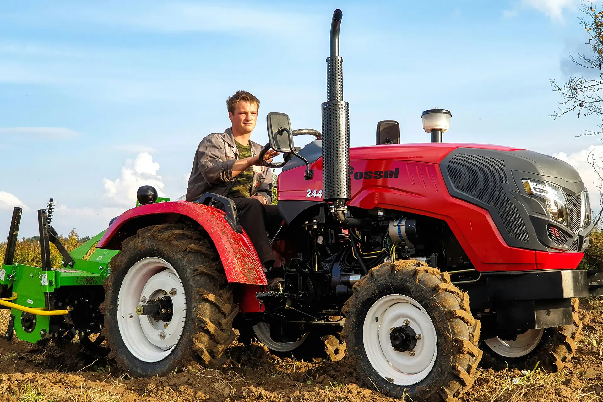 Kompakttraktoren eignen sich ideal für Arbeiten auf engem Raum und sind auch gebraucht eine attraktive Alternative. Junger Mann fährt einen roten Kleintraktor mit angebauter grüner Bodenfräse über einen frisch bearbeiteten Acker