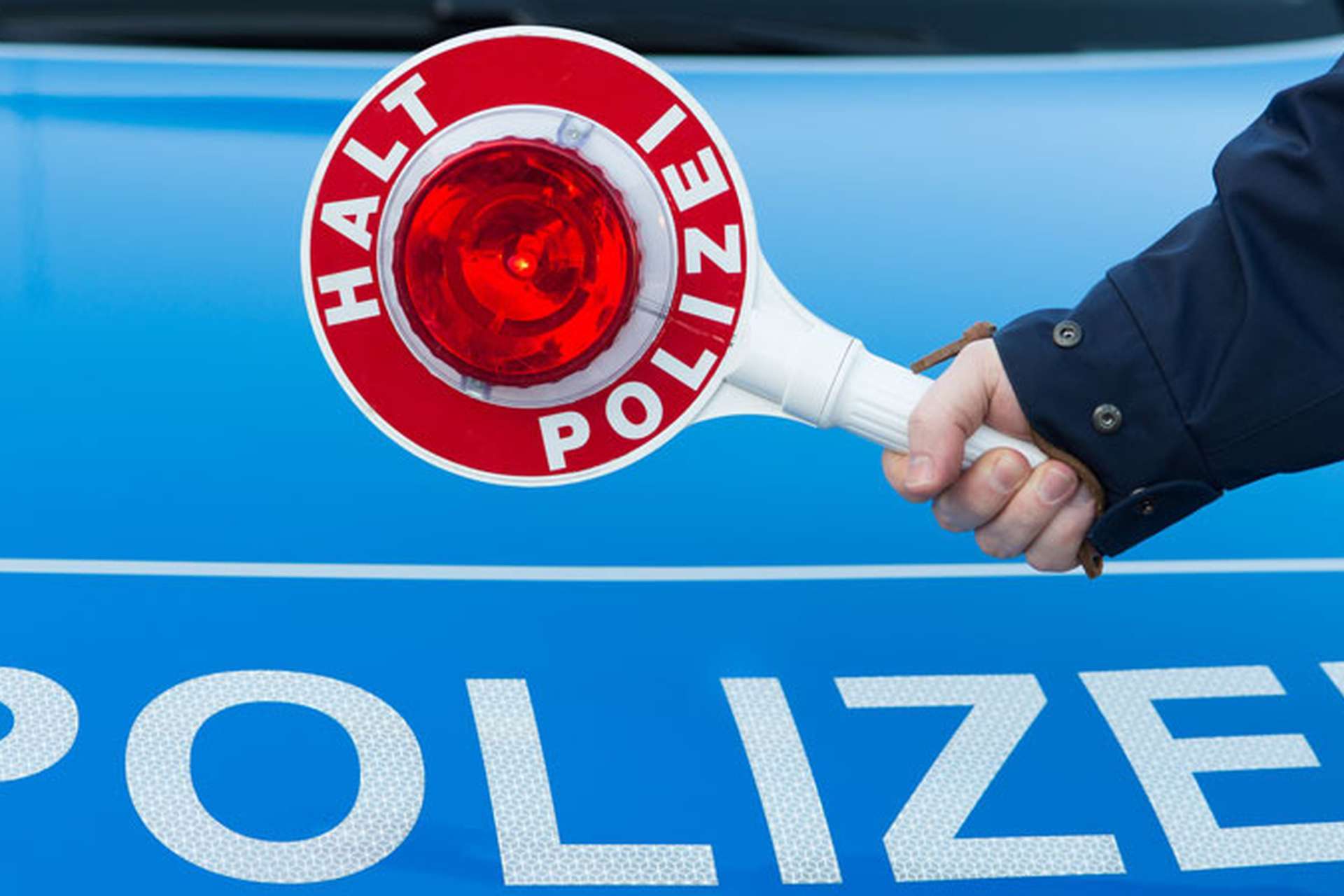 Arm der eine "HALT POLIZEI" Kelle hält mit Polizeischriftzug im Hintergrund